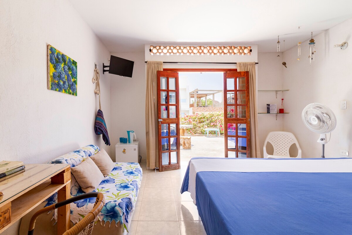 A well-lit room features a spacious bed covered with a blue bedspread. A sitting area with a floral-patterned sofa is present. Double wooden doors open to a view of a garden, enhancing the natural light. A television and a fan are mounted on the walls.