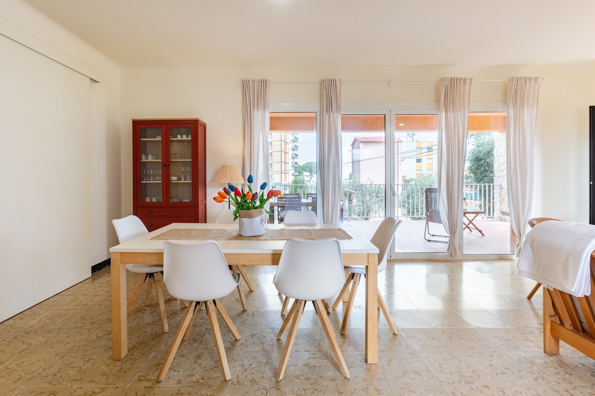 A spacious living area is furnished with a wooden dining table surrounded by white chairs. A vase of colorful flowers sits at the center. large glass doors lead to a terrace, allowing natural light to fill the room. Neutral tones create a calm environment.