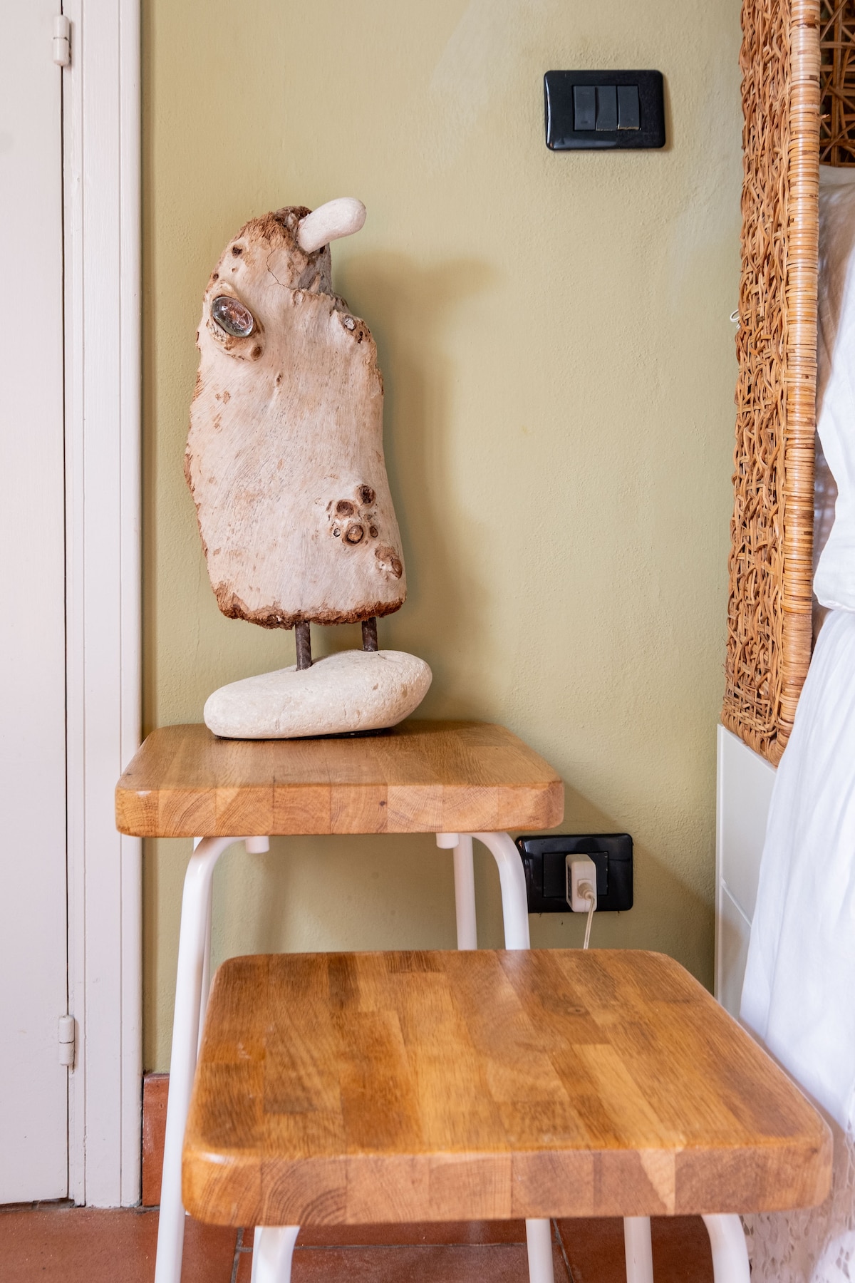 A simple wooden table stands beside a wall, supporting an artistic sculpture made from a gnarled piece of wood atop a smooth stone base. The room features neutral-colored walls, adding to the minimalist aesthetic.