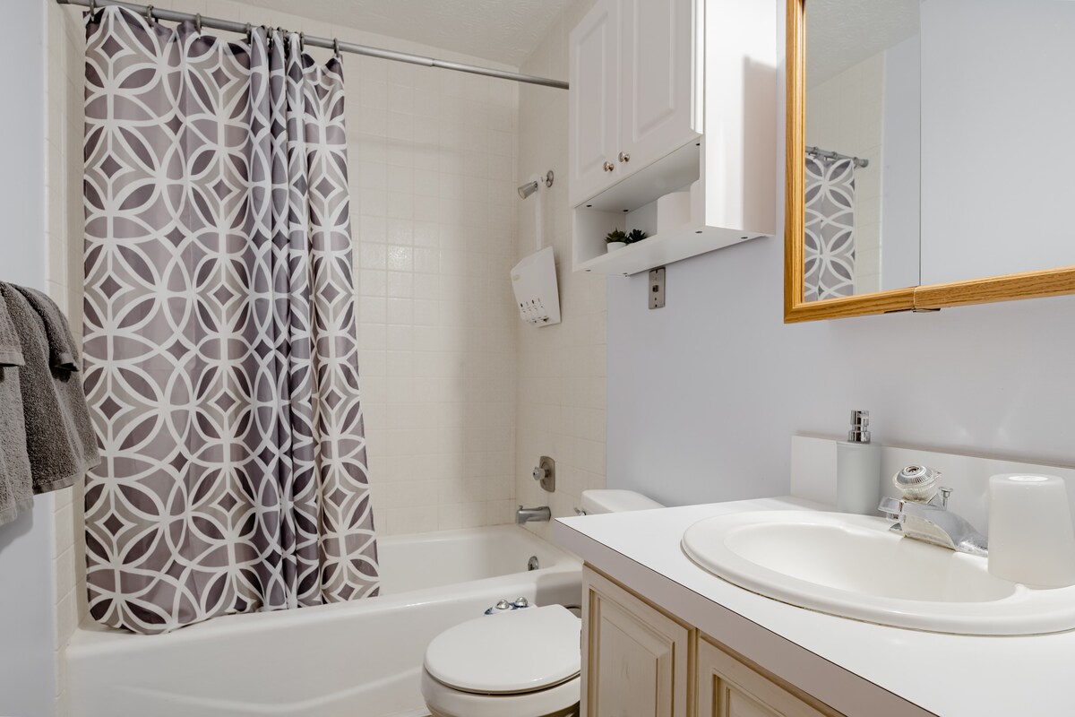 The bathroom features a combination tub and shower with a patterned curtain, and a light-colored countertop with a sink. A mirror is mounted above the sink. Storage options are available with cabinetry above the toilet and a shelf. Towels are hung on a rack.