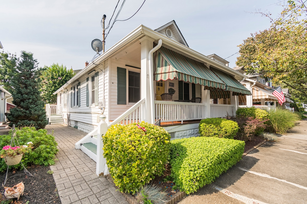 The charming exterior of the home is highlighted by a covered porch with striped awnings. Lush greenery surrounds the porch area, while a welcoming pathway leads to the front entrance. The property is set in a serene neighborhood, creating an inviting atmosphere.