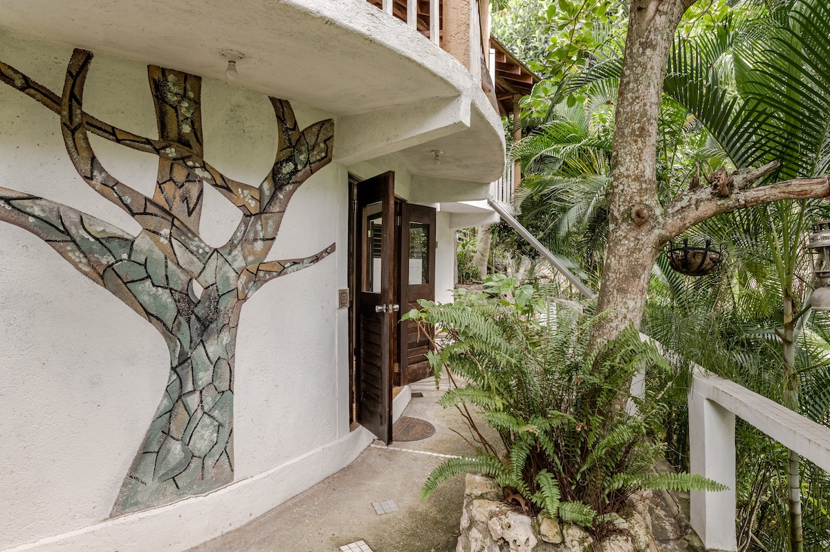 A unique entryway is framed by a decorative mosaic of a tree on the wall, surrounded by lush tropical plants. The curved structure features wooden doors, inviting guests into a serene natural setting. Pathways with stone accents guide the way through a vibrant garden.