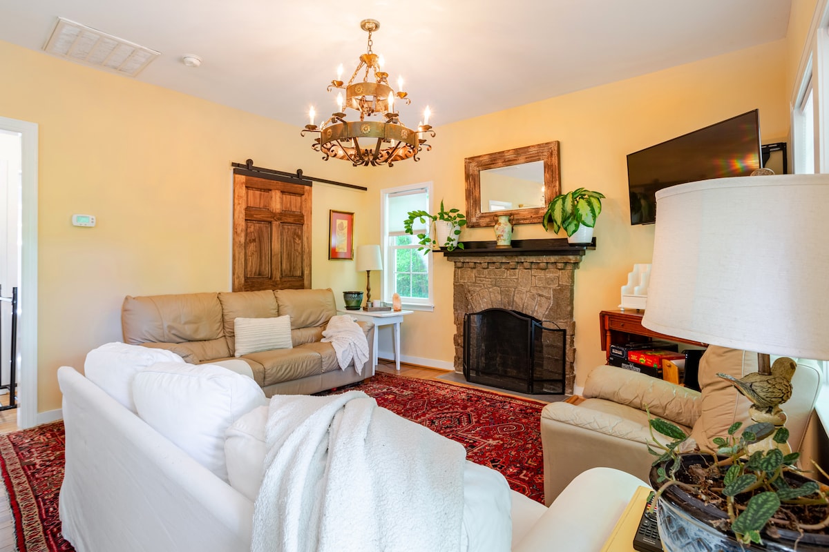A living area is presented with a neutral color palette, featuring a cream sofa and a light brown leather couch. Natural light illuminates the space through a window. A stone fireplace is flanked by a flat-screen TV and decorative plants, with a vintage chandelier overhead.