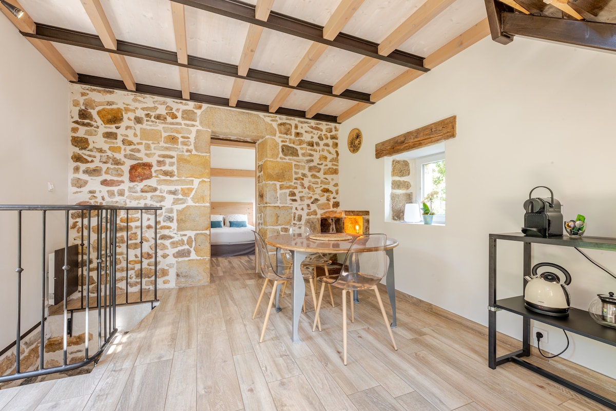 A charming living area showcases a round dining table surrounded by transparent chairs. Stone walls add character, while wooden beams are featured on the ceiling. A small kitchenette is visible, equipped with essential appliances. Natural light enters through a nearby window, enhancing the inviting space.