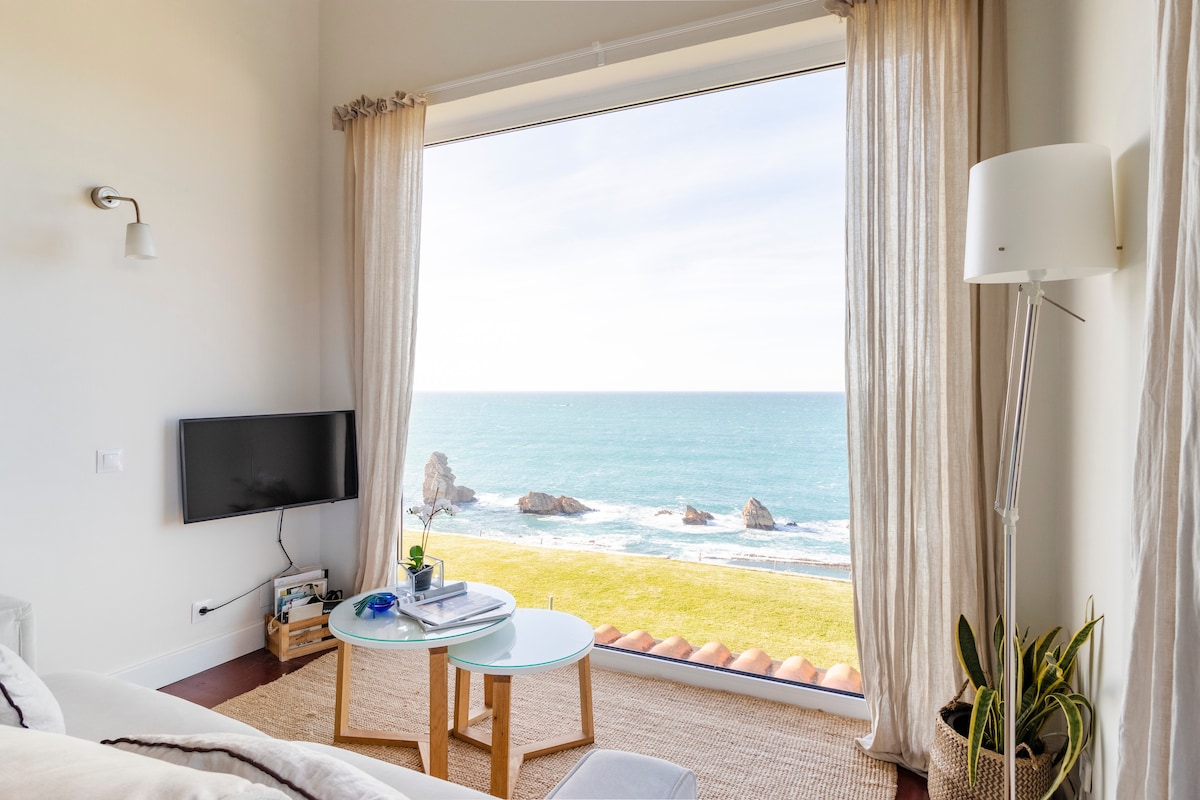 A living area features large glass windows that frame expansive views of the ocean and unique rock formations. A coffee table is placed on a woven rug, complemented by potted plants. Natural light fills the space, creating a serene ambiance.