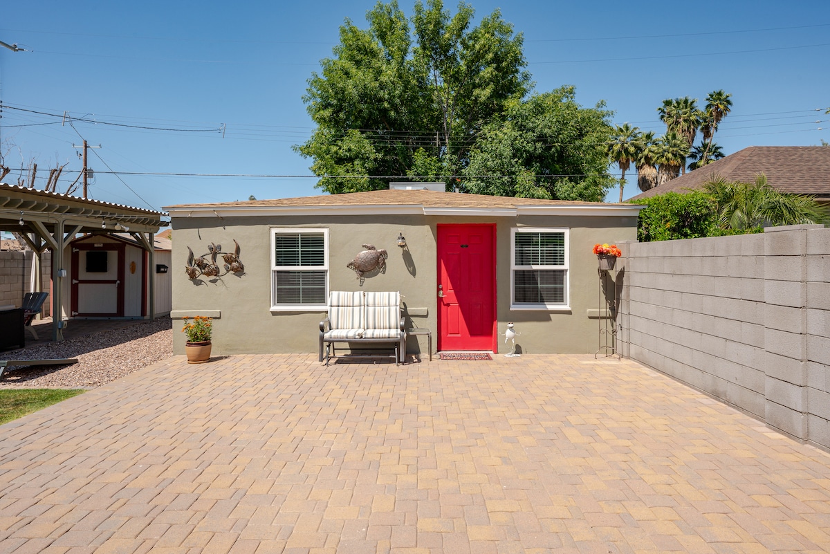 A single-story guest house is presented with a red front door and a comfortable white bench against a light gray exterior. The surrounding area features a paved stone patio and is complemented by lush greenery and palm trees in the background.