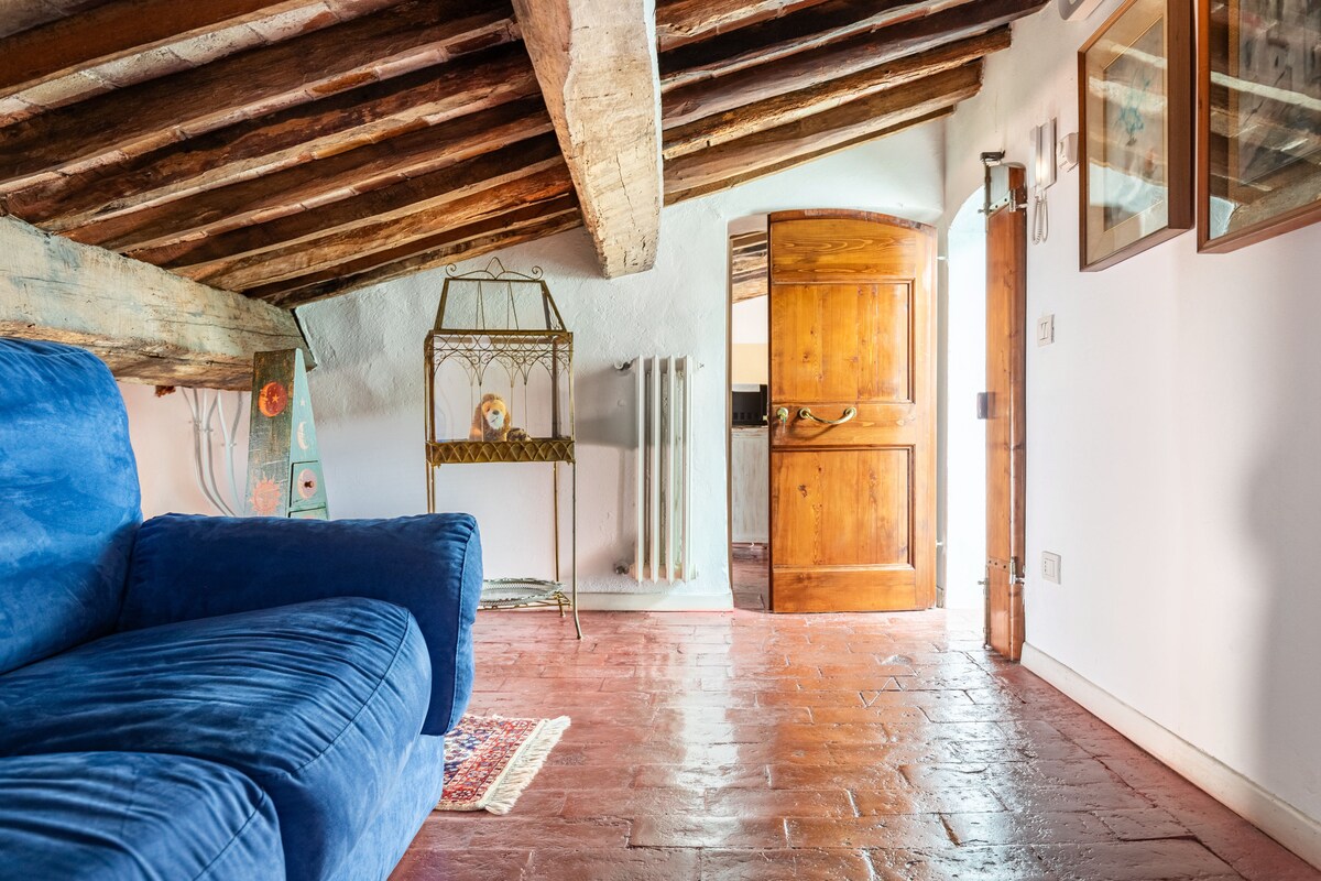 A cozy loft space features a plush blue sofa, exposed wooden beams overhead, and a warm terracotta floor. An inviting door opens to the exterior, with a decorative metal stand and wall art visible nearby, enhancing the room's character.