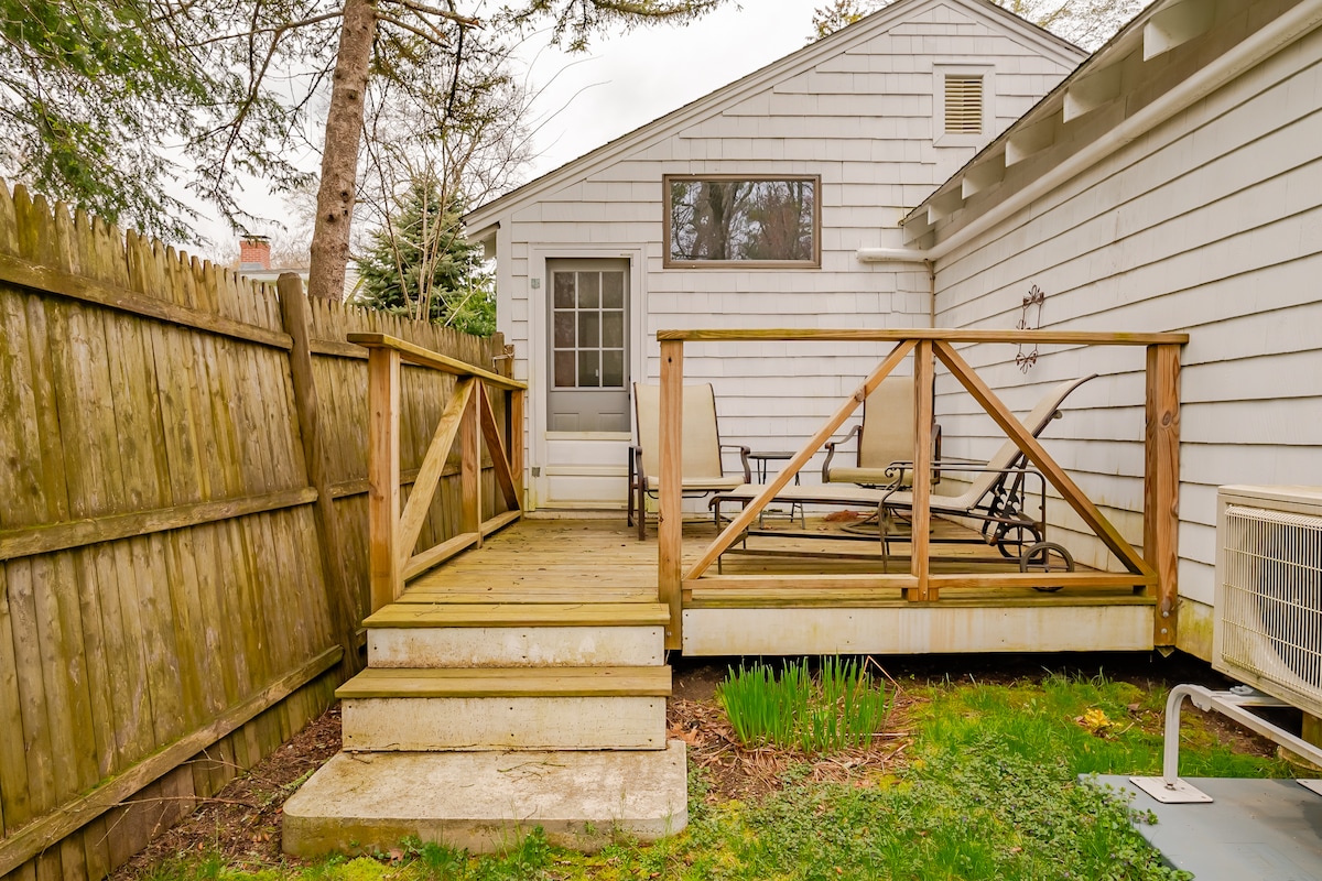 A private wooden deck is accessible via a small set of steps, featuring a railing for safety. The area is bordered by a wooden fence, and lounge chairs are arranged for relaxation. Green grass and shrubs surround the deck, enhancing the natural setting.