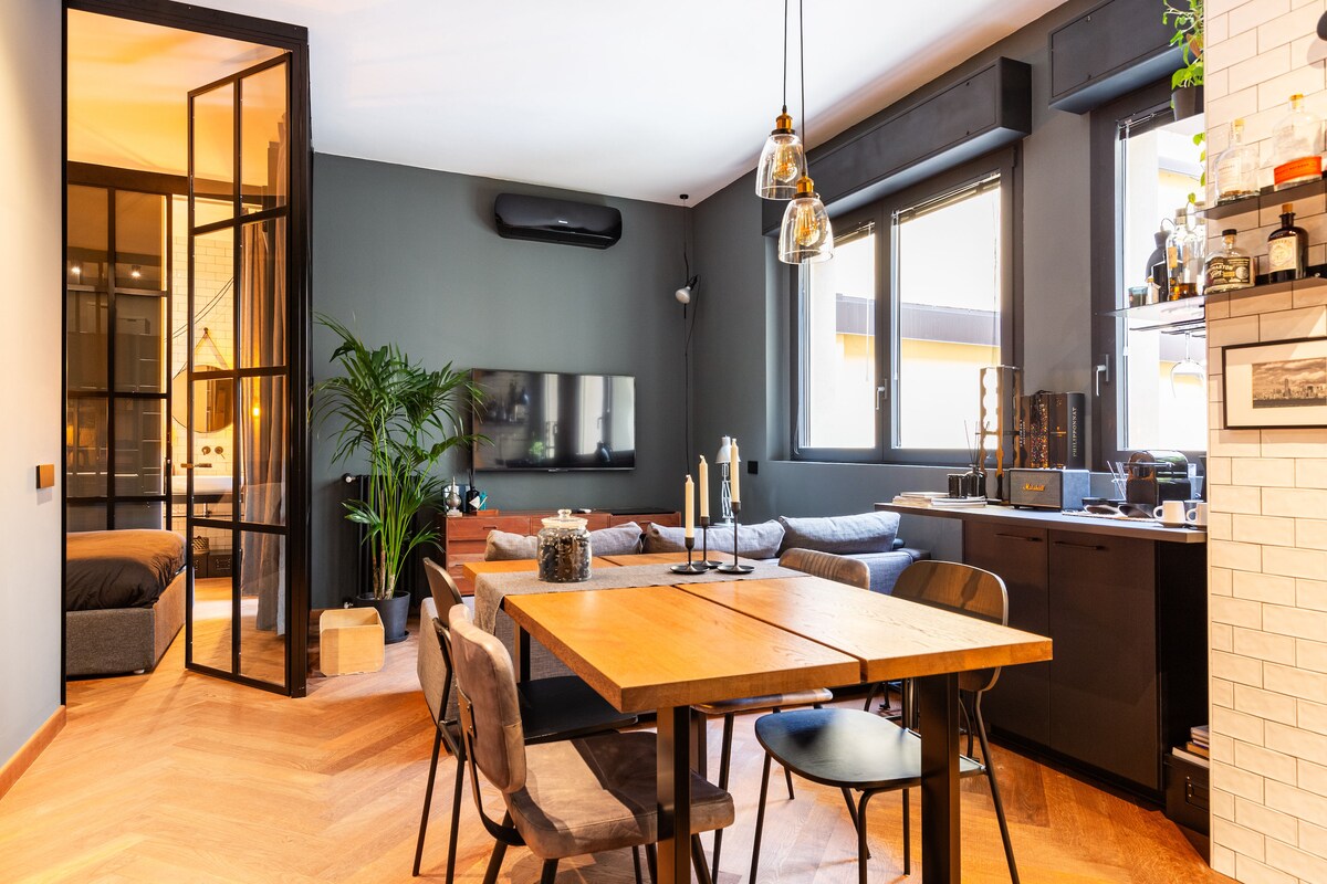 The living area features a wooden dining table surrounded by four chairs. A spacious sofa sits comfortably in front of a wall-mounted TV. Large windows permit natural light, and a glass partition separates the bedroom from the main space, enhancing the open design.