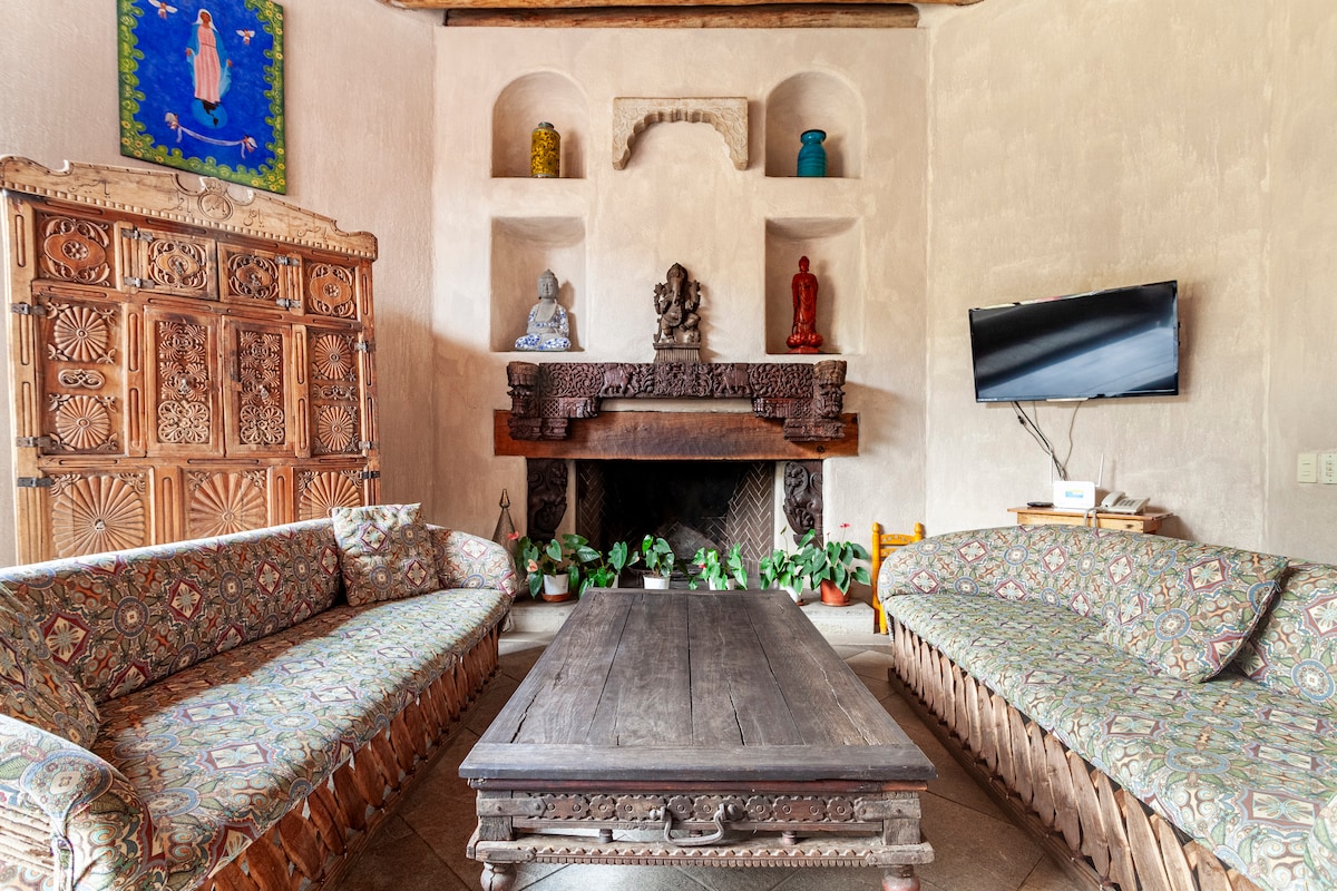 A spacious living area features two intricately patterned sofas flanking a wooden coffee table. The walls are adorned with decorative niches, housing colorful sculptures. A rustic fireplace sits below a carved wooden mantle, and a flat-screen TV is mounted on the wall.