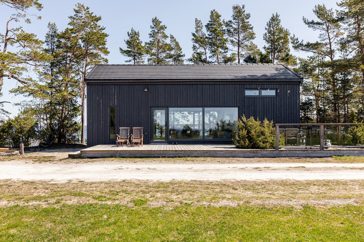 A modern cottage exterior is shown, featuring dark wooden walls and large glass windows that offer expansive views of the surrounding trees and landscape. A spacious deck with two wooden chairs and well-maintained greenery is visible, set against a clear sky.