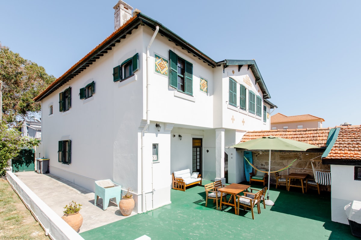 The two-story house features a spacious outdoor area with seating arrangements and a shaded dining space. Light-colored walls and dark green accents create a harmonious look. The surrounding greenery enhances the inviting environment, suitable for gatherings or relaxation.