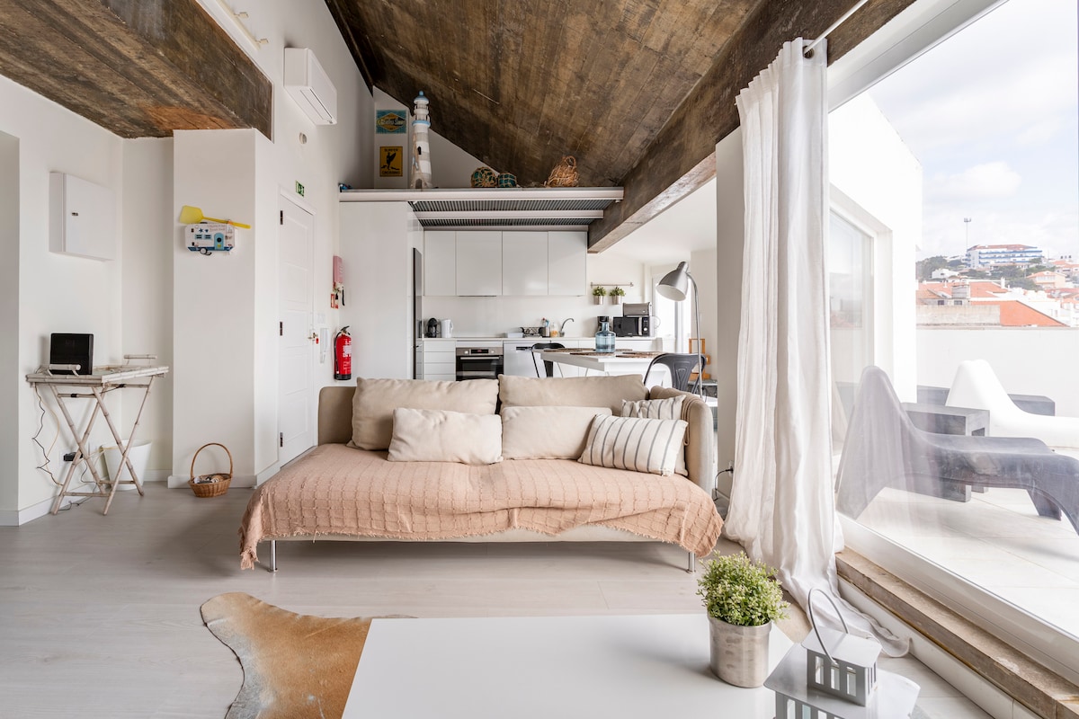 A modern living area is presented with high, wooden ceilings and a large window offering natural light. A light-colored sofa with a textured blanket is positioned in front of a stylish coffee table. The open kitchen area is visible in the background, featuring sleek cabinetry and appliances.