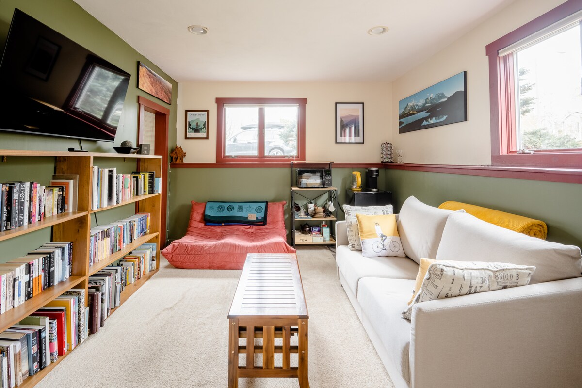 A cozy living area is presented with a beige sofa and a wooden coffee table. A red beanbag chair provides additional seating, while a bookshelf filled with various books stands against the wall. Natural light enters through two windows, enhancing the room's welcoming vibe.