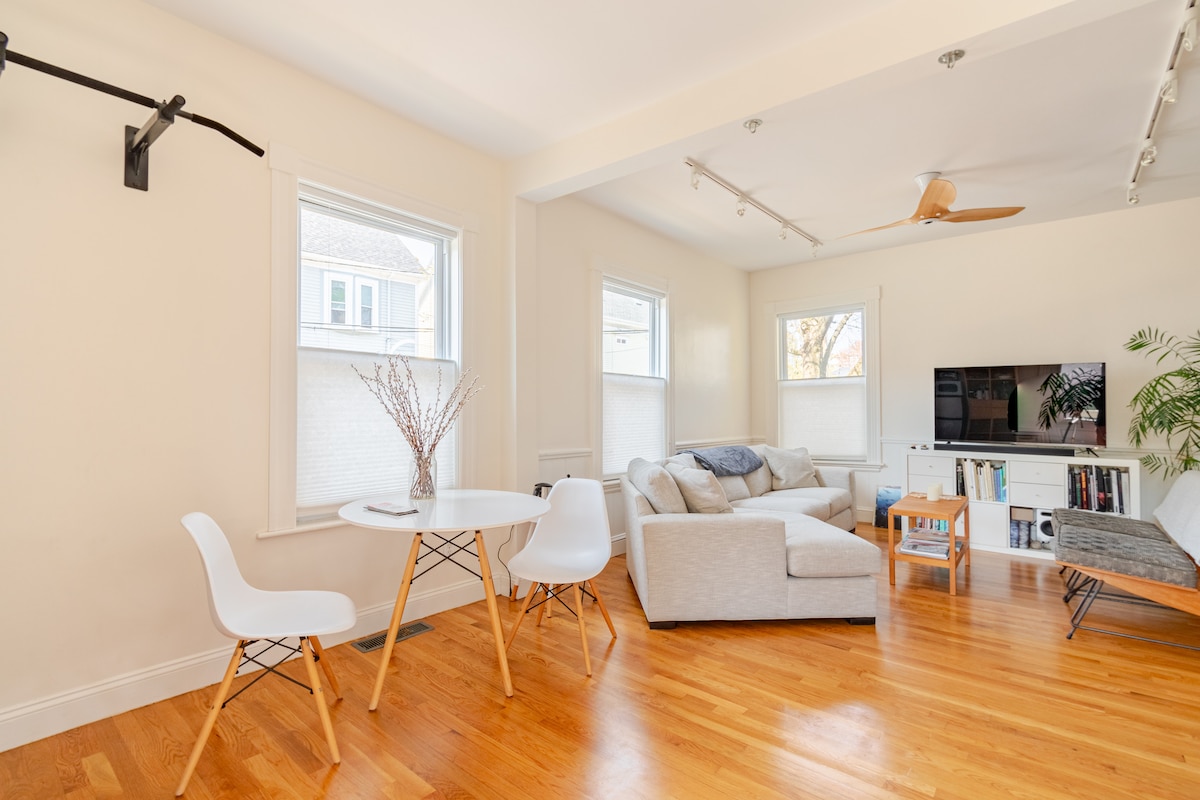 The living room features light wood flooring and an open layout. A mix of seating options includes a gray sectional sofa and a minimalist chair set around a small white table. Natural light streams through two windows, highlighting the peaceful interior.