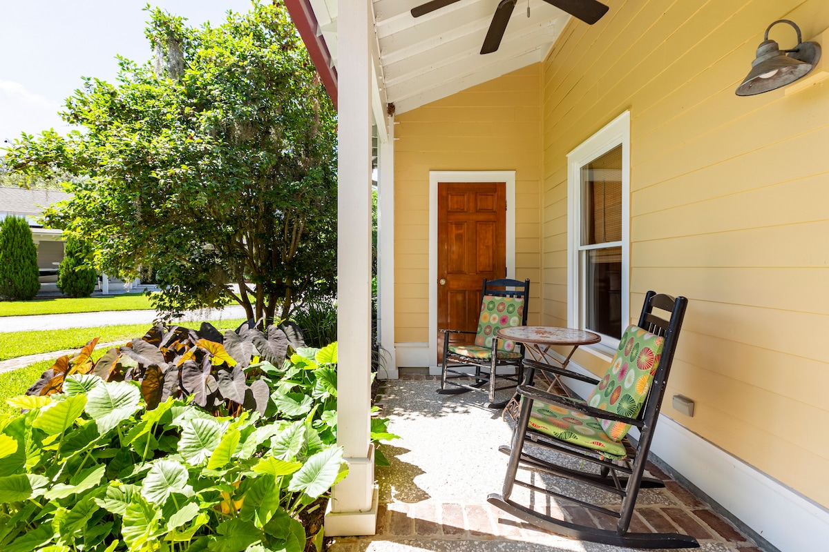 A charming porch area is showcased, featuring two rocking chairs with colorful cushions positioned around a small table. Lush green foliage and ornamental stones frame the space, while a wooden door offers an inviting entrance. Natural light illuminates the inviting outdoor area.