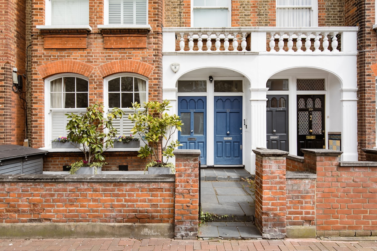 The image displays a charming row of brick buildings featuring two blue front doors, with decorative white architraves. Lush green plants are positioned near the entrance, while a stone pathway leads to the doors. Large windows with white frames provide natural light.