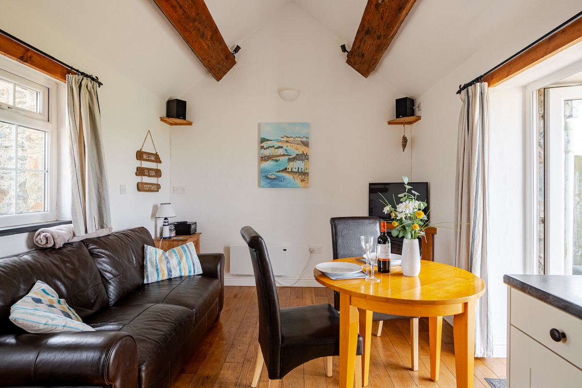 An open-plan living area showcases a comfortable leather sofa complemented by decorative cushions. A wooden dining table is set for two, surrounded by natural light from patio doors. A vibrant painting hangs above, contributing to the home's welcoming ambiance.