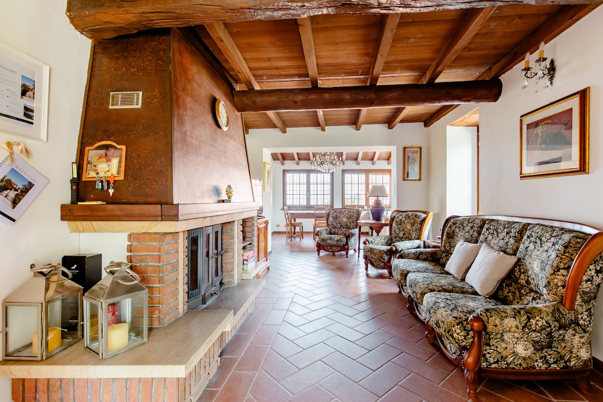 A welcoming living area features classic furnishings, including floral-patterned sofas and armchairs. A brick fireplace serves as a focal point, while a wooden beam ceiling adds character. Natural light enters through large windows, with a dining area visible in the background.