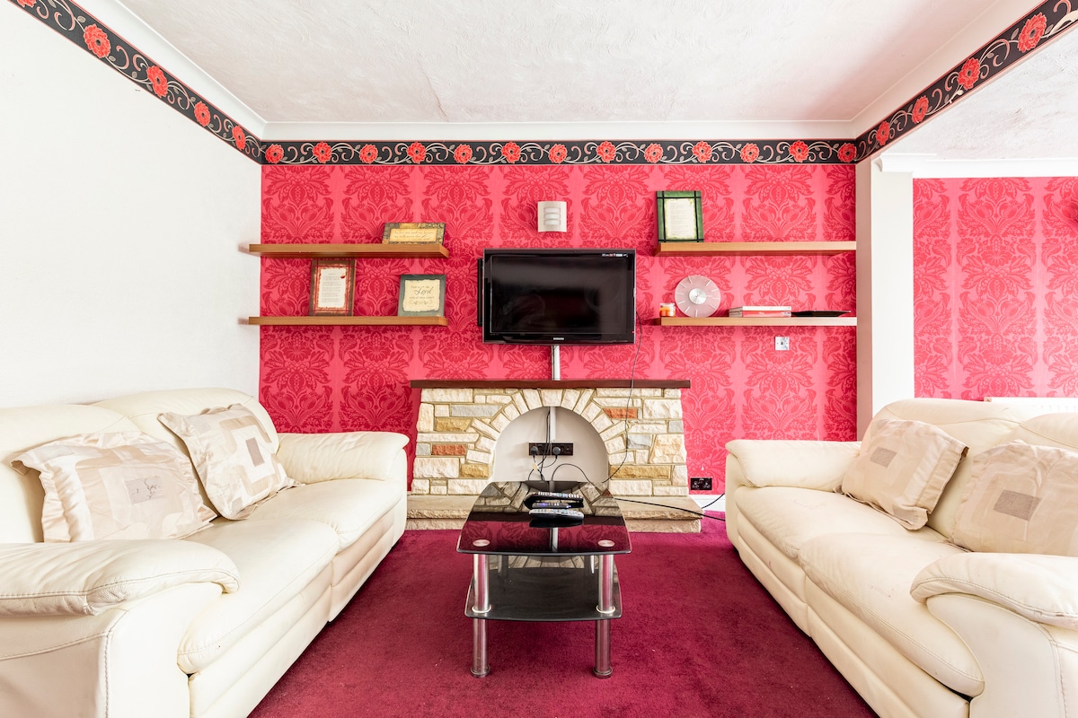 A living area features two cream-colored sofas positioned parallel to each other, with a glass coffee table in between. A flat-screen television is mounted on a wall with decorative shelving above. The backdrop is adorned with bold red wallpaper, complemented by a stone fireplace.
