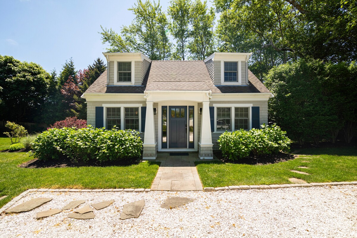 The exterior of a charming cape-style house is presented, featuring a well-maintained yard with green shrubbery and a gravel pathway. Two dormer windows complement the front, while a welcoming door is flanked by elegant lanterns, surrounded by lush trees providing shade.