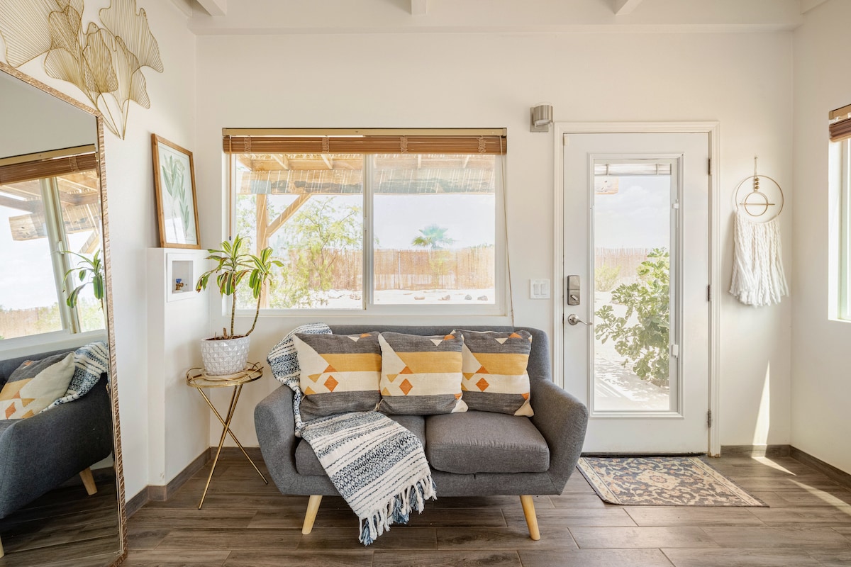 A cozy seating area features a gray sofa adorned with patterned cushions, positioned near large windows that allow natural light to fill the space. A stylish mirror reflects the room, while a decorative plant adds a touch of greenery. The entrance door leads to the outside.