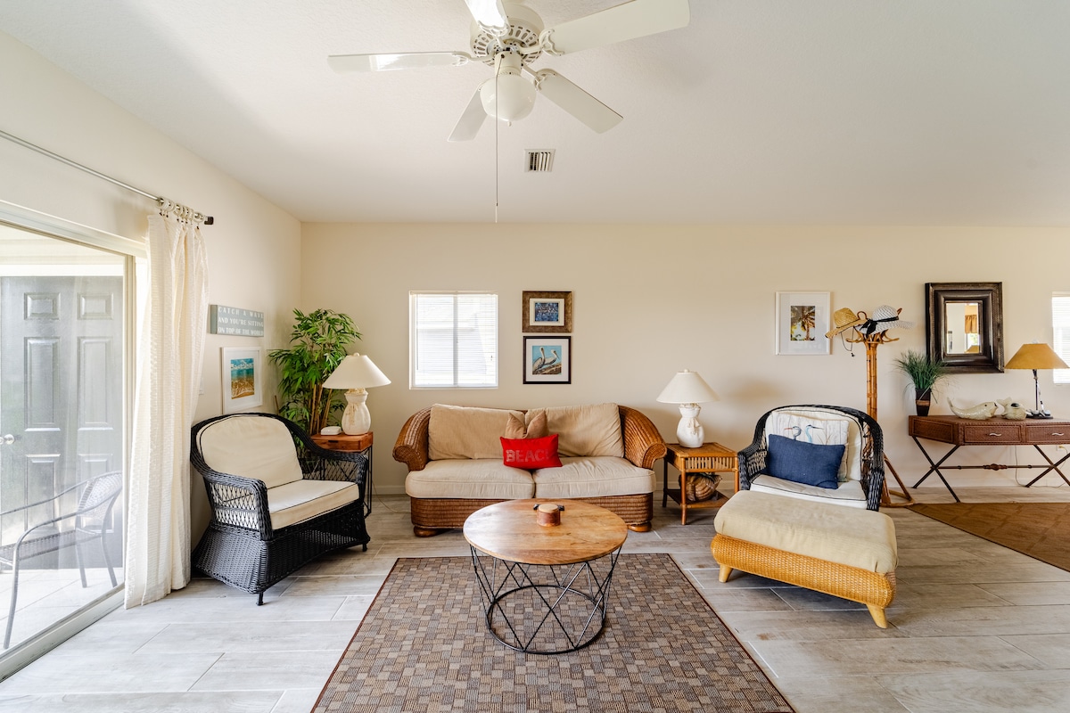 A spacious living area is furnished with a mix of wicker and fabric seating, including a dark wicker chair and a light chaise lounge. A round coffee table anchors the space, while light enters through windows with minimal coverings. The room is bright and inviting, featuring neutral tones.