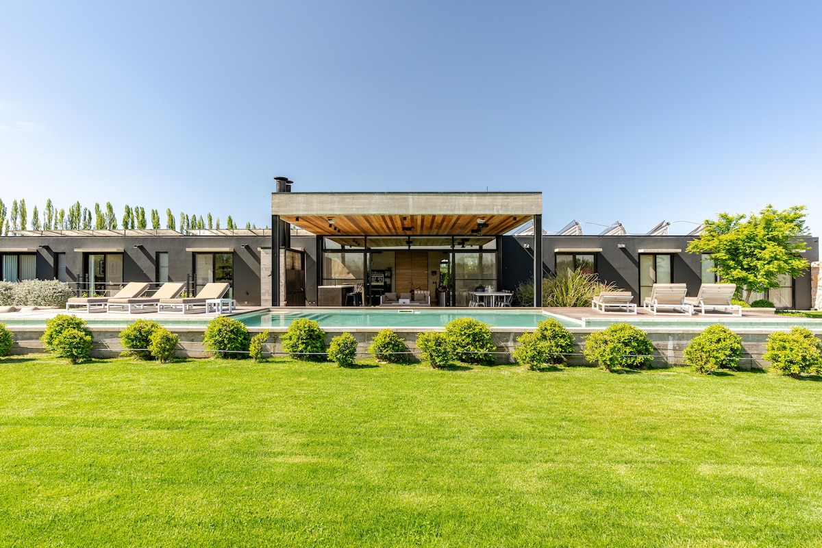The modern exterior of Casa Viña is showcased, featuring a spacious terrace that overlooks a swimming pool surrounded by well-maintained grass. Sun loungers are positioned around the pool, and tall trees line the background, creating a serene and open atmosphere.