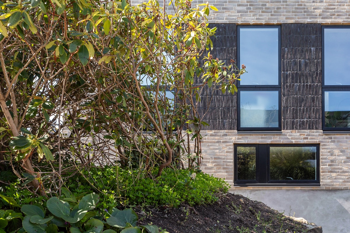 The exterior of the building features large, symmetrical windows framed in black, set against a backdrop of light-colored brick. A lush, green shrubbery surrounds the base, adding a natural element to the modern architecture.