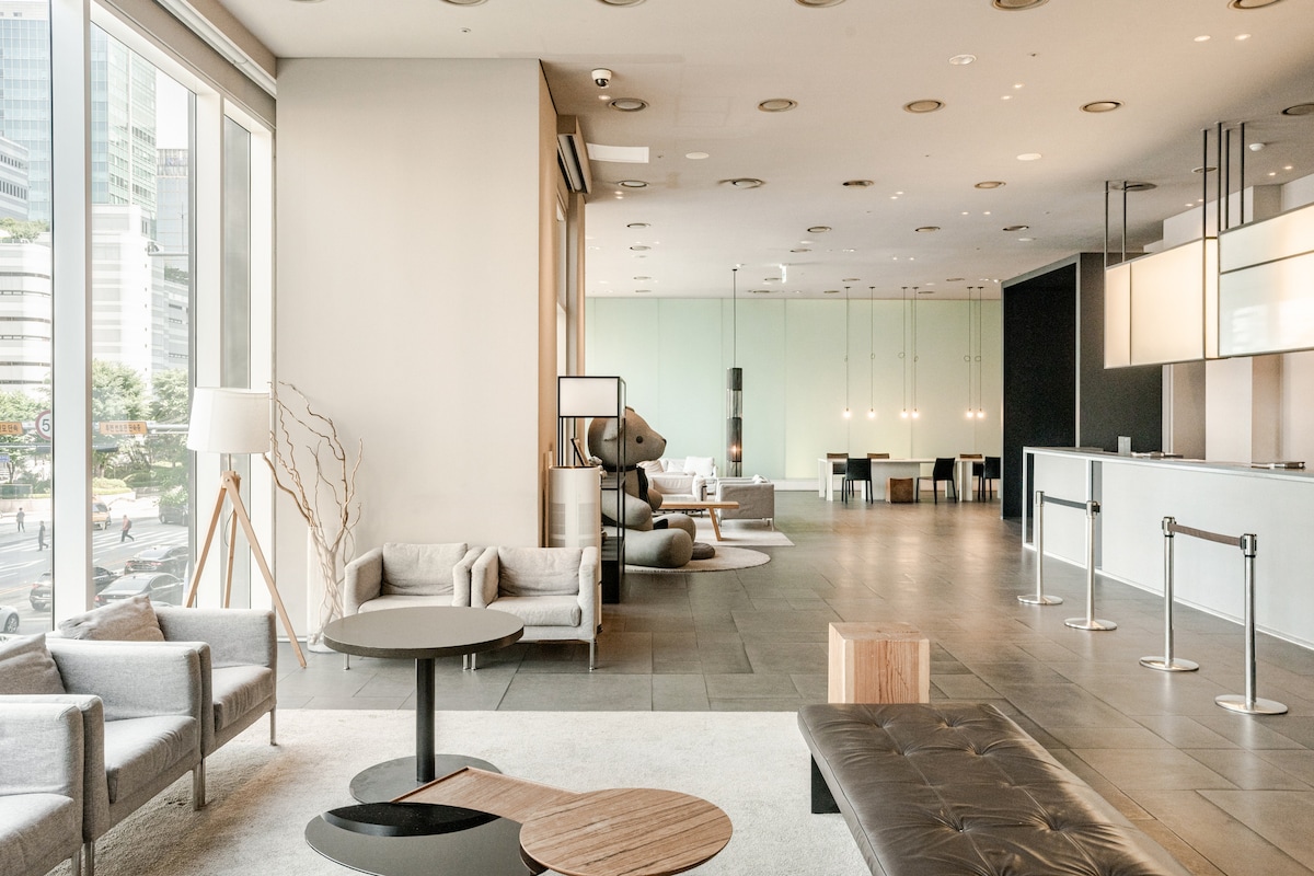 A modern lobby area features a spacious layout with sleek furniture. Soft grey couches and seating arrangements are complemented by minimalistic tables. Large windows provide ample natural light, and the reception area is designed with a contemporary counter, enhancing the overall modern aesthetic.