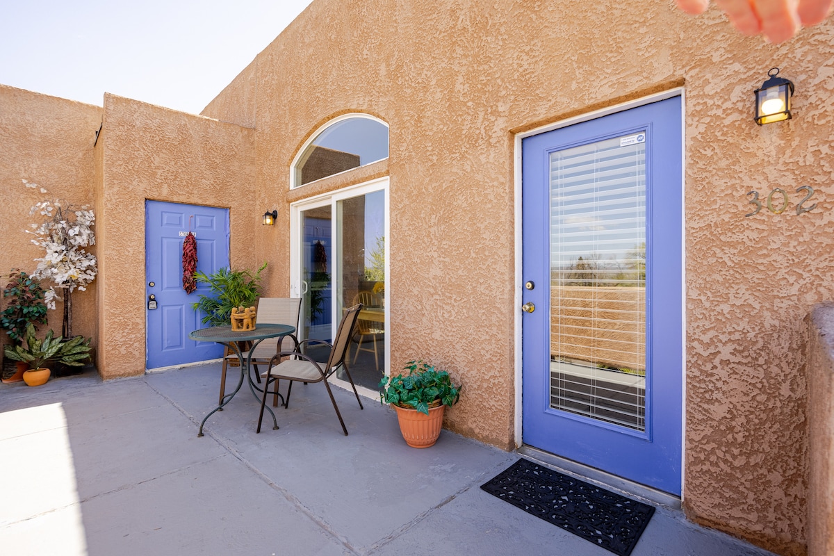 The entrance to a light-colored building is visible, featuring arched windows and two blue doors. A small table with two chairs sits on a patio, accompanied by potted plants. Sunlight illuminates the area, highlighting the textured wall and inviting outdoor space.