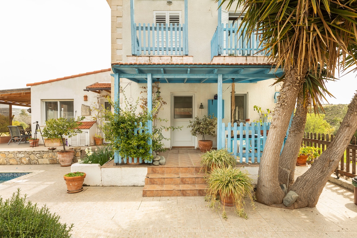 The exterior of the house features a welcoming entrance with terracotta tiles and a blue wooden railing. Green plants in pots are placed around the area, and a glimpse of the swimming pool is visible to the left. A shaded terrace is situated toward the back.