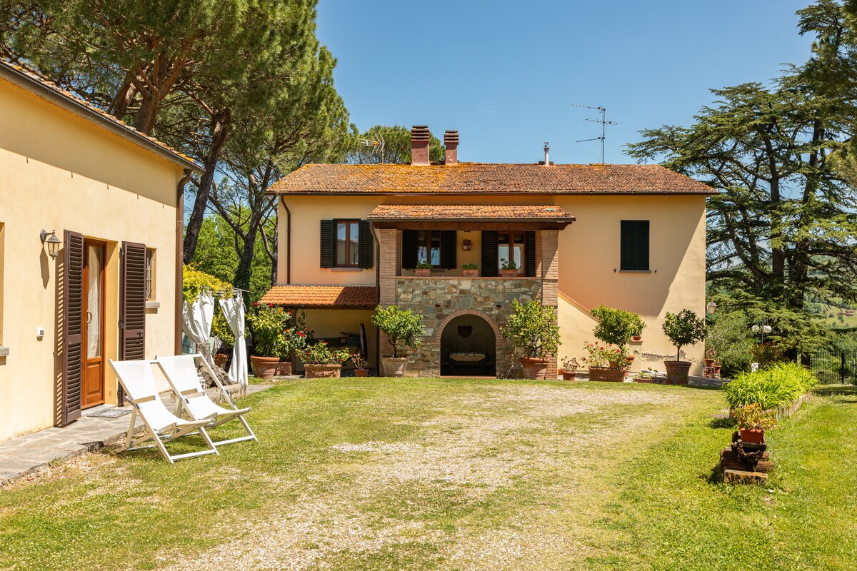A charming two-story residence is set within a spacious garden, featuring a welcoming entrance with stone accents. Potted plants and neatly arranged outdoor seating enhance the serene environment. Sunlight bathes the area, highlighting the harmonious blend of nature and architecture.