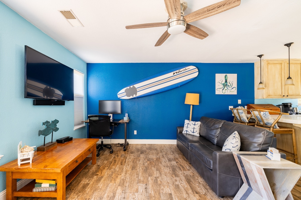 A living area features blue accent walls with a surfboard mounted decoratively. A spacious dark sofa is placed opposite a wooden coffee table. A flat-screen TV is mounted on the wall, while light streams in through a window with partially closed blinds.