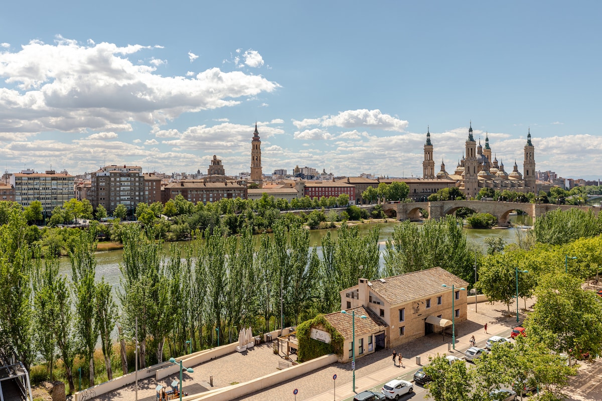 A panoramic view captures a river lined with lush greenery and several buildings in the background, including a majestic cathedral and other structures, under a partly cloudy sky, providing a vibrant contrast to the tranquil water below.