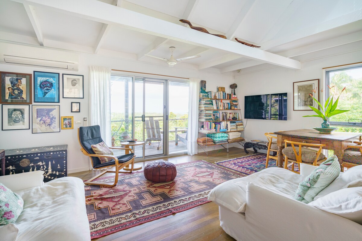 An open-plan living room is displayed with large glass doors leading to a balcony, inviting natural light. A rocking chair and a decorative pouf are positioned on a patterned rug. Shelves filled with books and a green plant add to the inviting atmosphere.