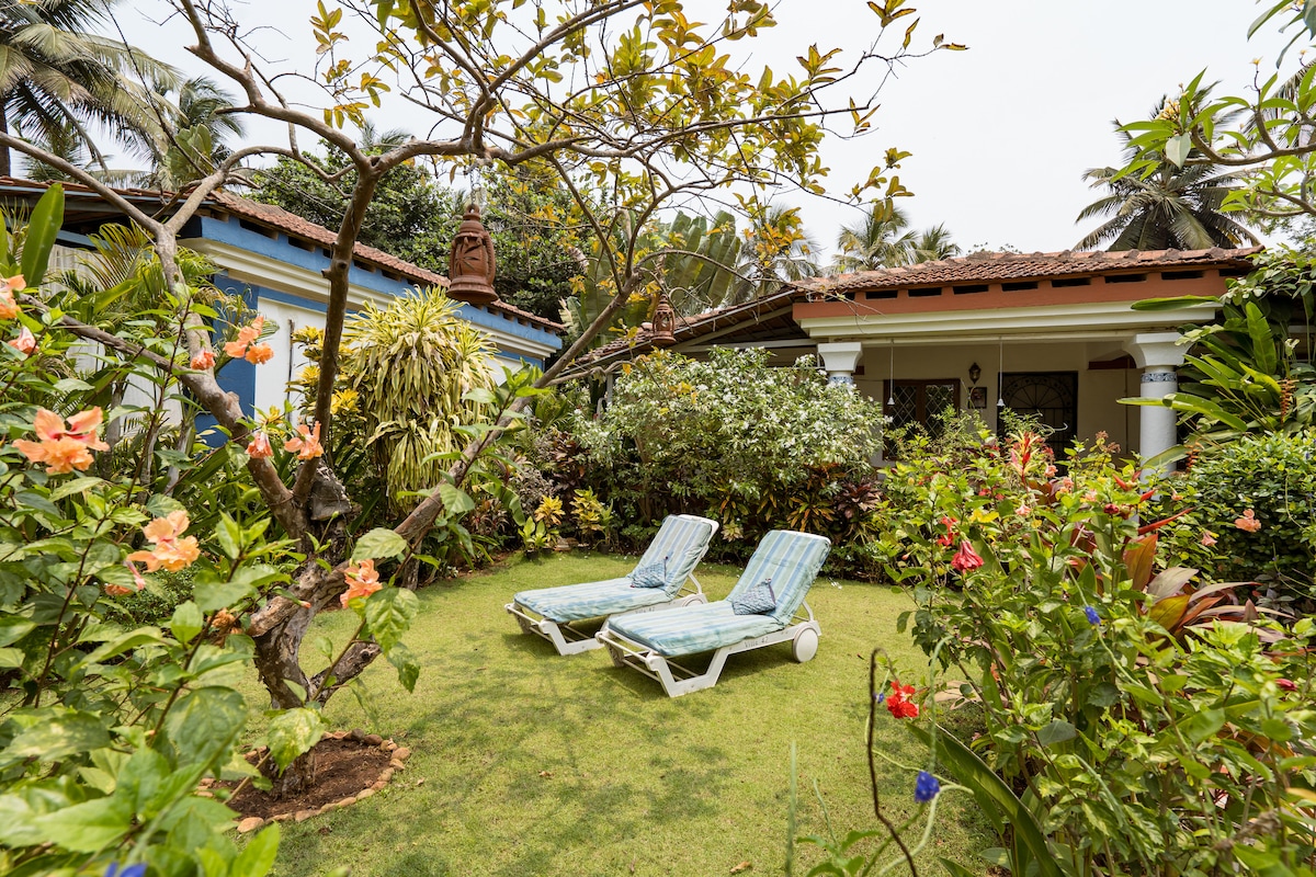 A serene garden area is depicted with two lounge chairs positioned on a lush green lawn. Surrounding tropical plants and colorful flowers contribute to a tranquil environment, while the villa's patio and roof are visible in the background under a clear sky.