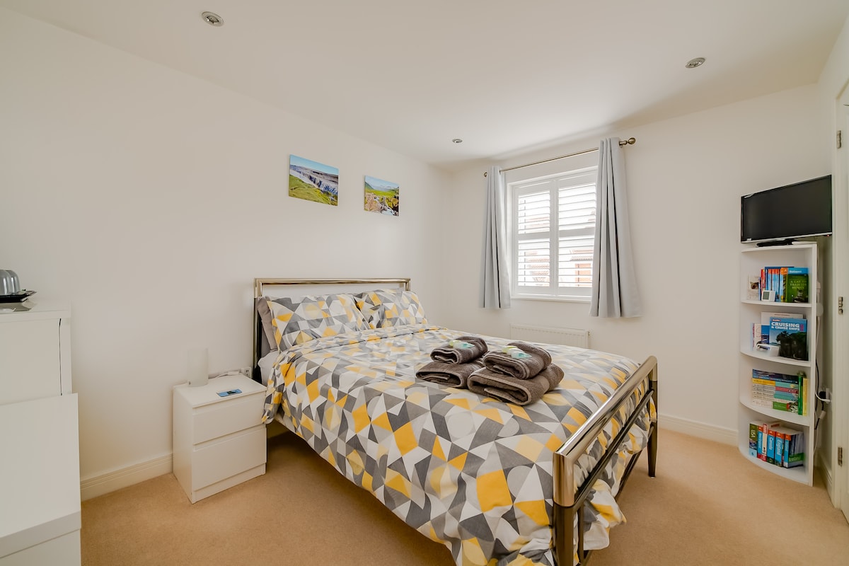The bright bedroom features a spacious bed adorned with a geometric patterned duvet cover. Natural light enters through a window with blinds. A TV is mounted on the wall, and a bookshelf filled with various titles is present in one corner. The decor is simple and clean.