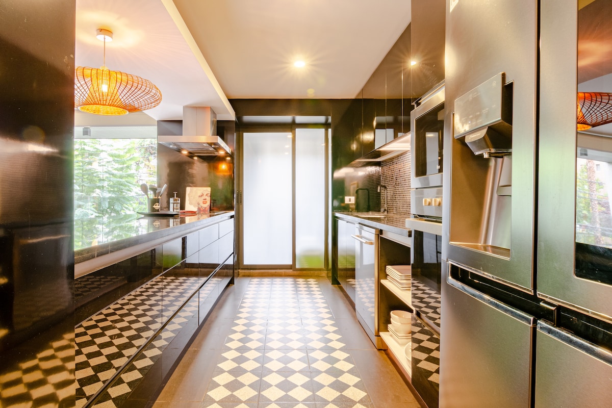 A spacious kitchen with dark cabinetry and modern appliances is seen, featuring a side-by-side refrigerator, oven, and dishwasher. The black and white tiled floor creates a geometric pattern, while natural light enters through a large window, enhancing the layout.