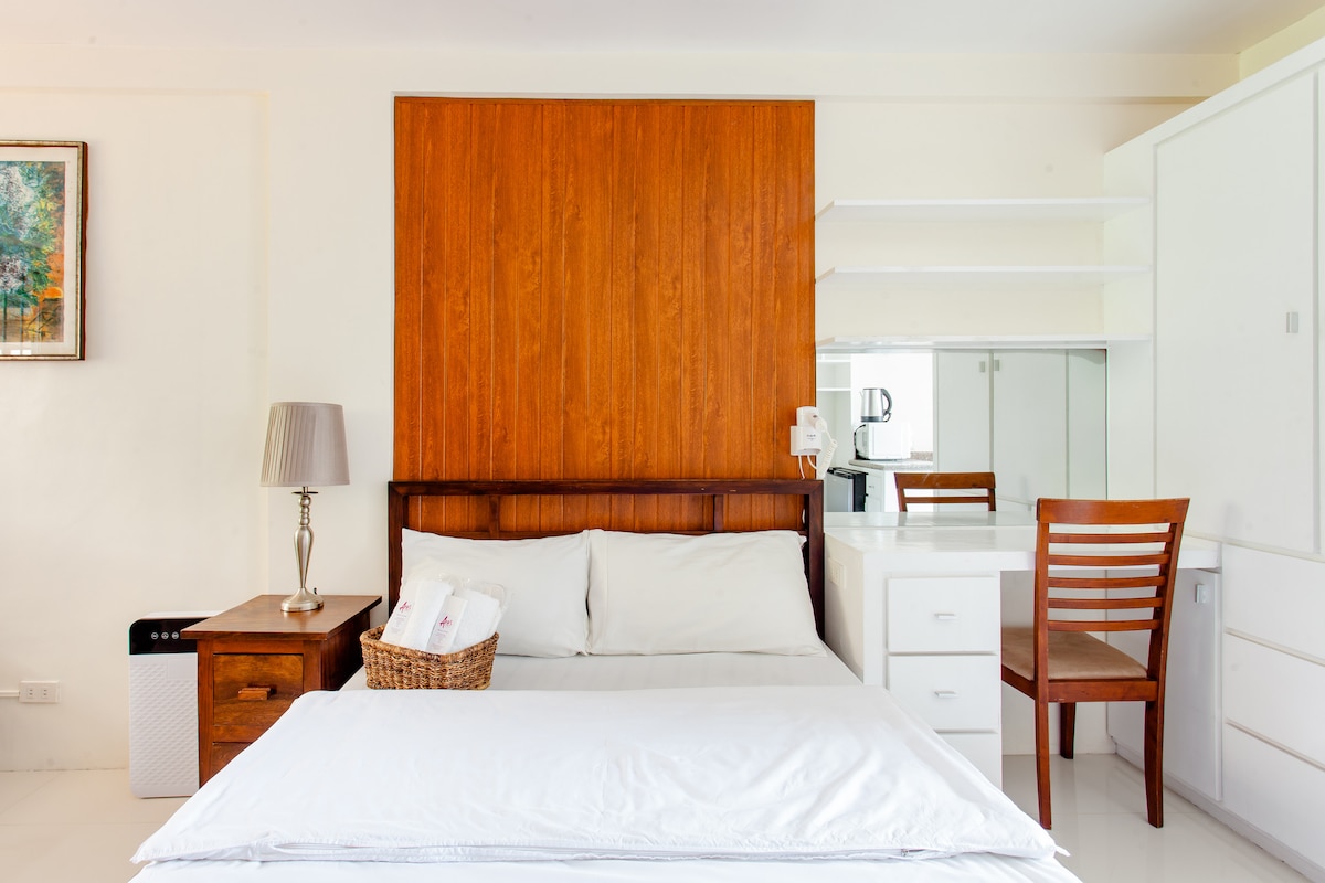 A well-appointed studio room features a spacious bed with neutral linens, complemented by a wooden headboard. A bedside table with a lamp and a wicker basket sits next to the bed. A wooden chair and storage cabinets are visible, providing essential amenities.