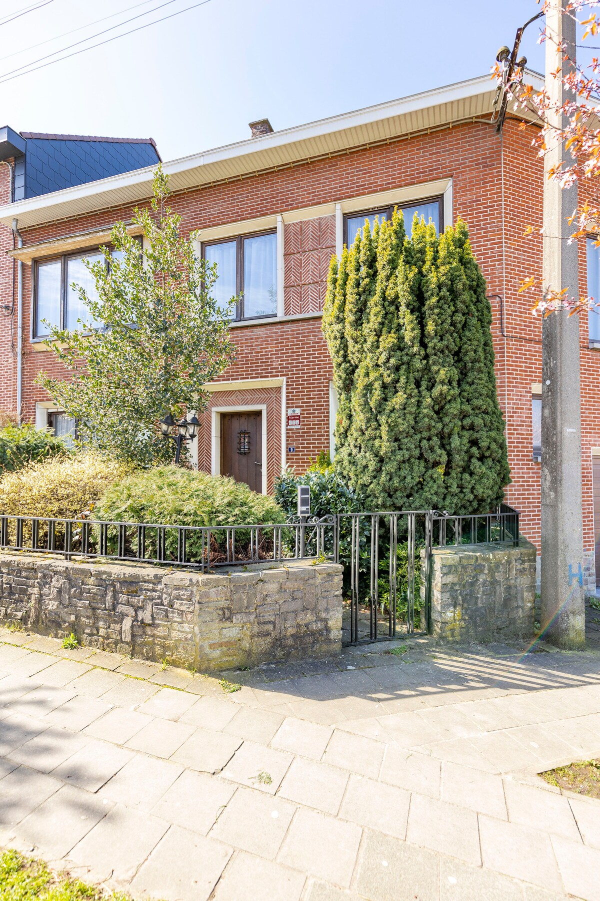 A brick-faced house is seen from the street, featuring large windows framed by green shrubs and a tall evergreen tree. A stone wall and wrought iron gate mark the entrance, creating a welcoming facade. Clear blue skies provide a bright backdrop.