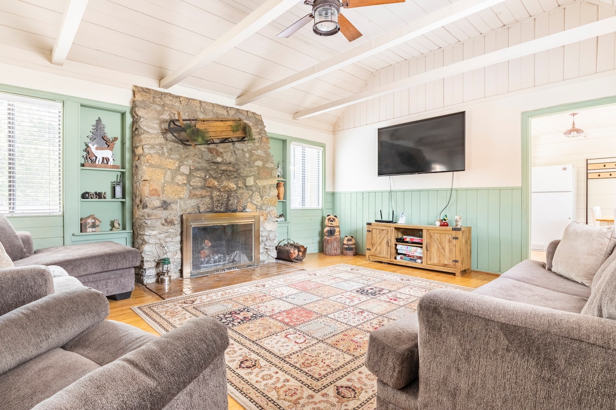 A comfortable living space features a stone fireplace as the focal point, with a large screen television mounted above it. Two cozy sofas are arranged around a patterned area rug, creating a welcoming area for relaxation and social interaction. Natural light filters through the windows.