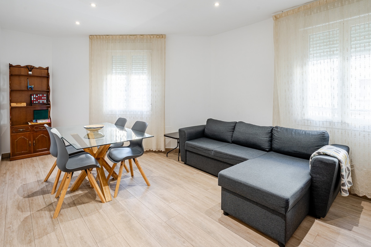 The living area features a comfortable gray sectional sofa and a modern dining table with six chairs. Natural light fills the space through large windows, complemented by light, sheer curtains. A wooden cabinet is visible in the background, adding functional storage.