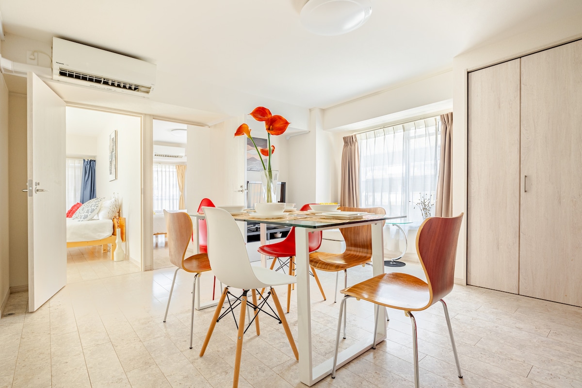 A dining area features a round glass table surrounded by six chairs in various colors. Natural light illuminates the space through large windows with light curtains. An air conditioning unit hangs above, while an open doorway leads to a hallway.