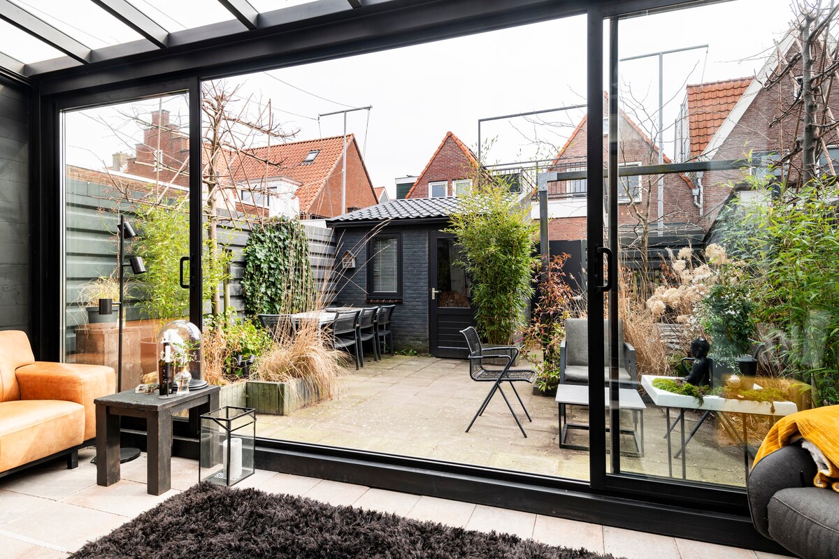 A view of the private enclosed garden is presented, with greenery and seating options visible. A sleek black table and chairs are arranged for outdoor dining, resting on stone paving. The garden is complemented by a cozy lounge area, surrounded by various plants and garden features.