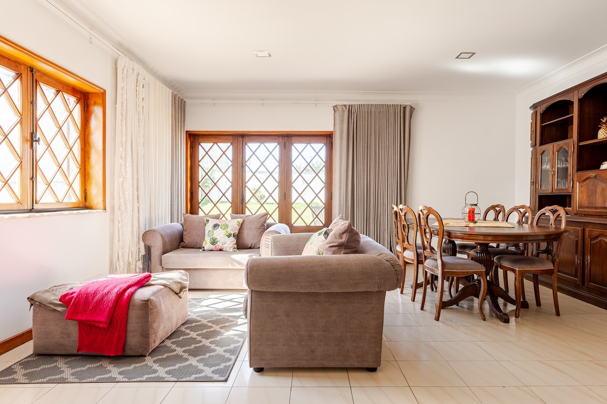 A living area is composed of a comfortable sofa and chair, accented by decorative cushions. Large windows with lattice designs allow natural light to fill the space, which is complemented by a dining table with wooden chairs, surrounded by soft curtains and a patterned rug.