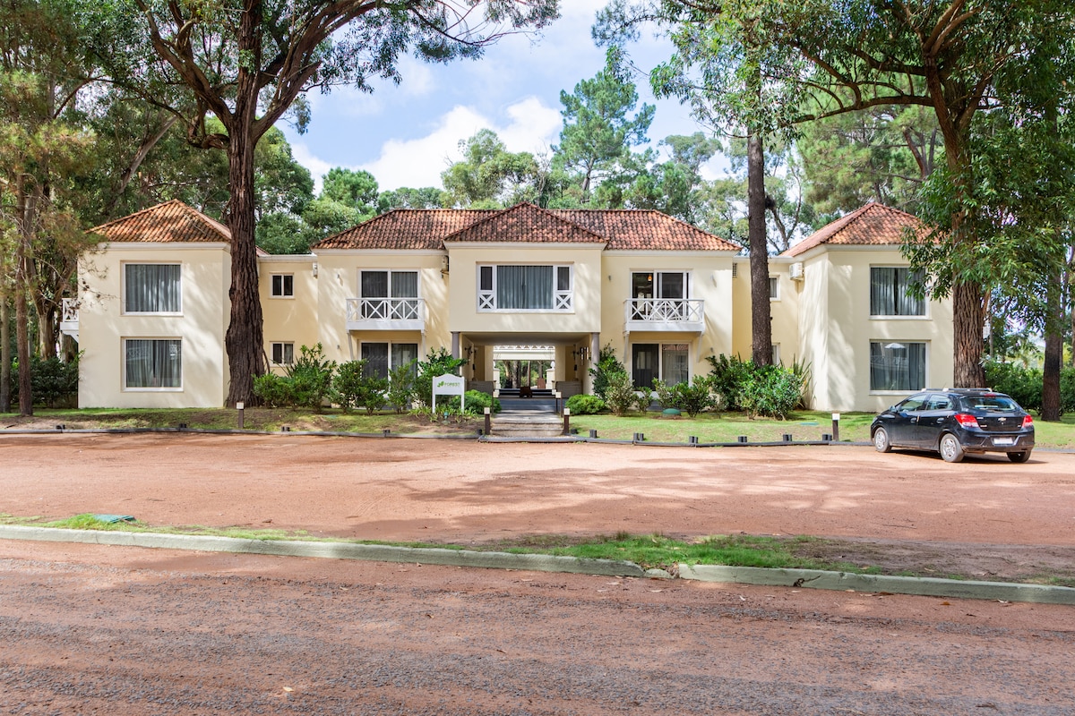 A multi-story building is set among tall trees, featuring a symmetrical design with a mix of stone and light-colored walls. Large windows bring in natural light. The entrance is marked by a set of steps leading to a welcoming doorway, while a parked car is visible in front.
