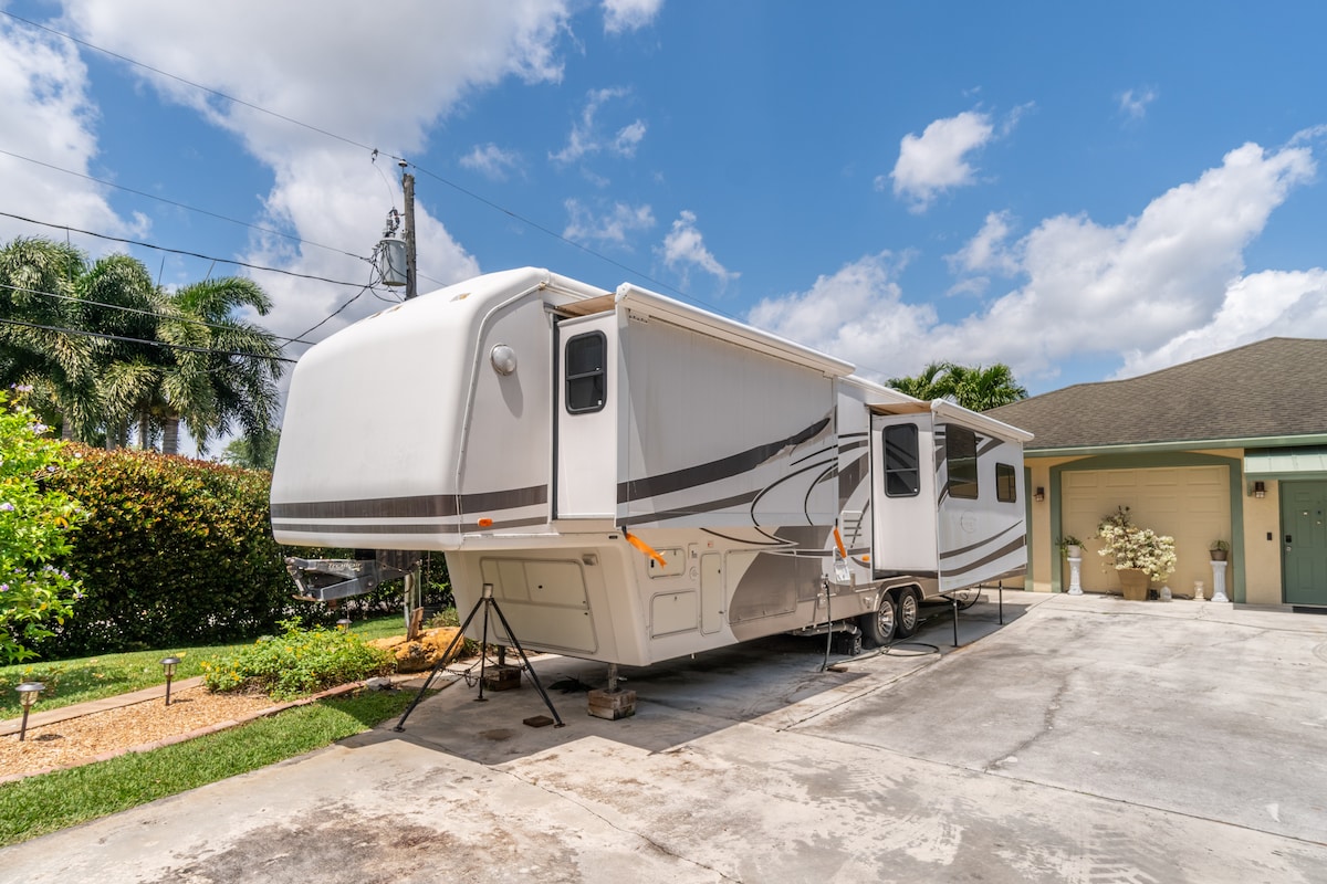 The RV is parked on a concrete driveway, featuring a streamlined design and modern exterior. Surrounding tropical plants are visible, contributing to a serene environment. The sky is partly cloudy, with hints of blue peeking through, enhancing the inviting atmosphere of the setting.