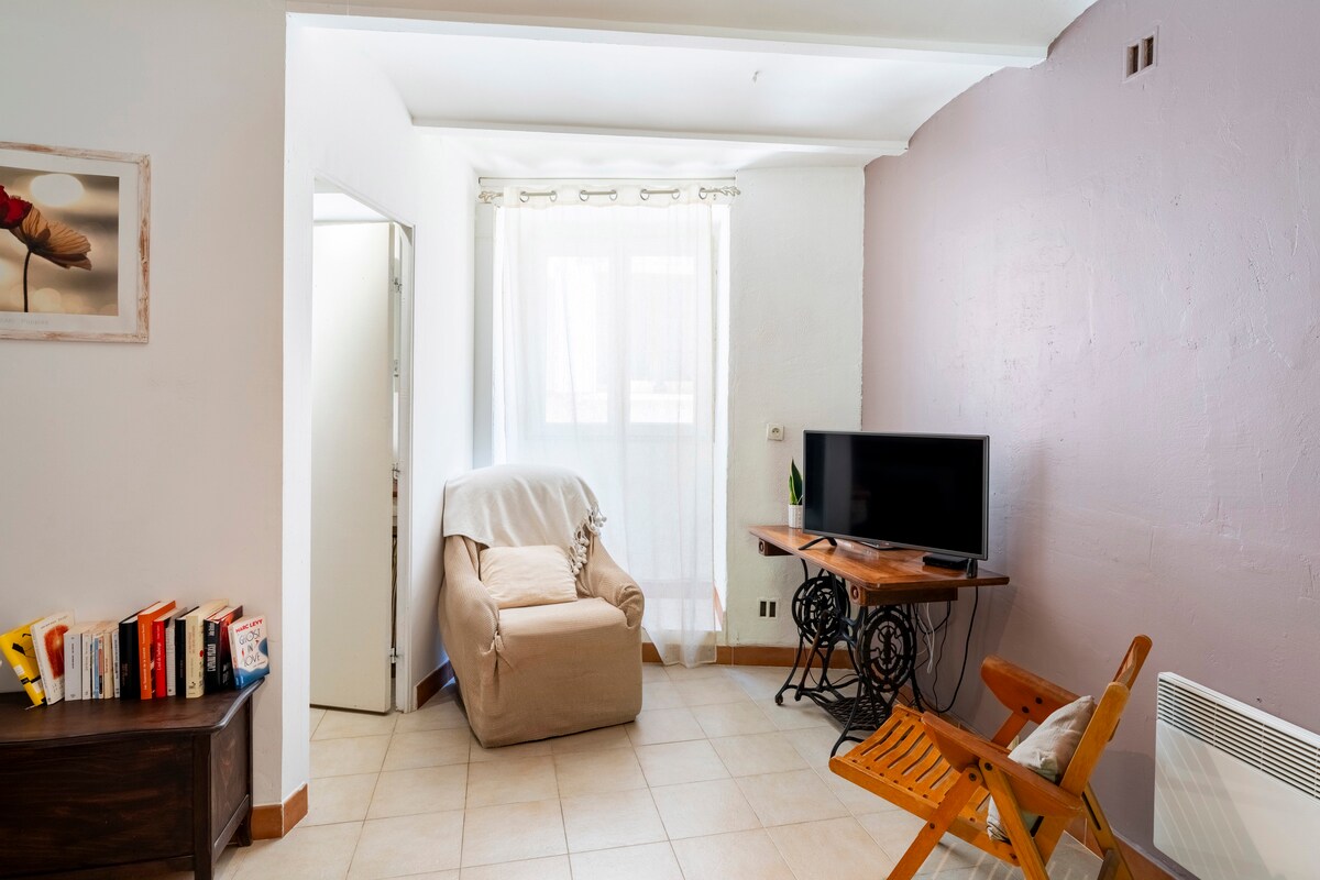 A comfortable seating area is provided with a soft armchair and a wooden folding chair. Light filters through sheer curtains, illuminating the space. A wooden table supports a television, and a selection of books is arranged on a nearby shelf, enhancing the cozy environment.