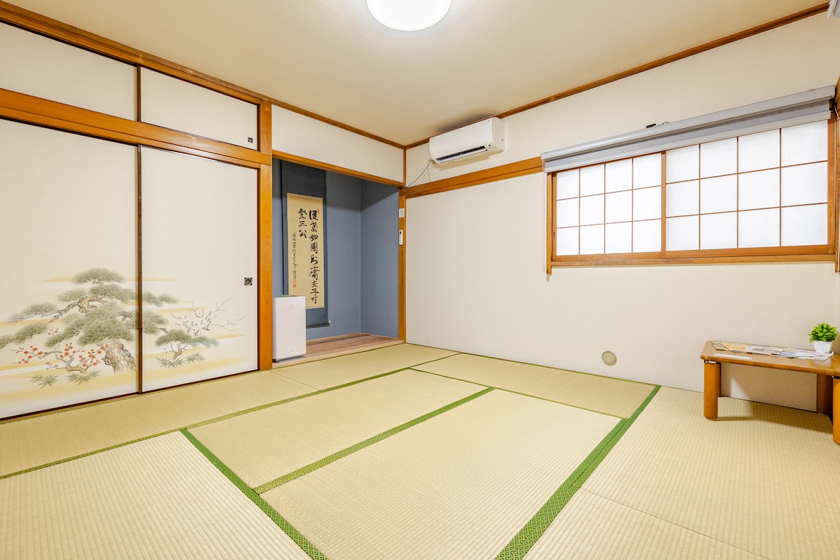 A traditional tatami room features sliding doors with an intricate design. Natural light fills the space through a window adorned with a simple grid pattern. A small table is set against one wall, and the room provides a clean, spacious environment suitable for relaxation.
